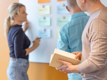 Mann hält Notizbuch in Hand. Im Hintergrund arbeiten Kollegen an Whiteboard.