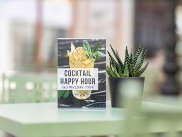 Tischaufsteller mit Cocktail Happy Hour steht auf Tisch neben Pflanze