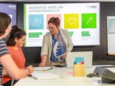 Frau steht vor Leinwand und gibt Kollegin eine Erklärung