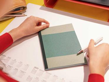 Frau im roten Pulli sitzt am Schreibtisch mit blauem Notizbuch auf Papier-Schreibunterlage