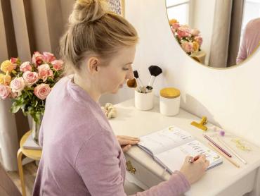 Frau in rosa Pulli sitzt Zuhause an Schreibtisch und notiert in Notizbuch, Blumenstrauß im Hintergrund