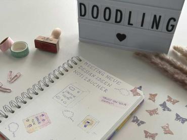 Notizbuch mit Doodles liegt auf Tisch, oberhalb steht das Wort "Doodling" auf einer Anzeigentafel, daneben Heftklammern und weitere kleinere Accessoires