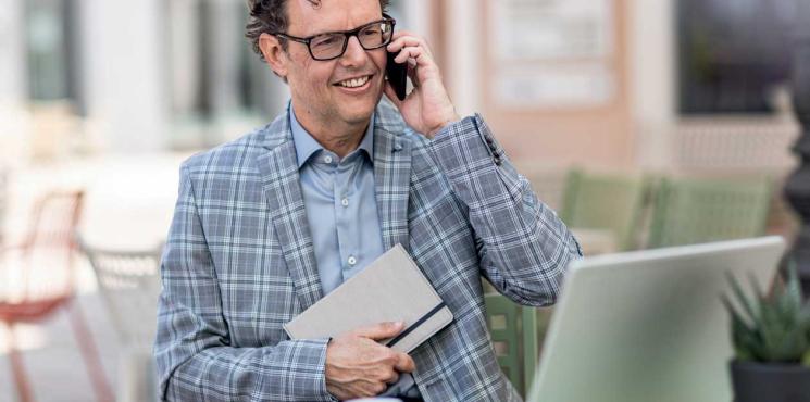 Mann sitzt mit Laptop und Notizbuch im Café und telefoniert