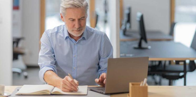 Business-Mann notiert in Notizbuch, Laptop daneben