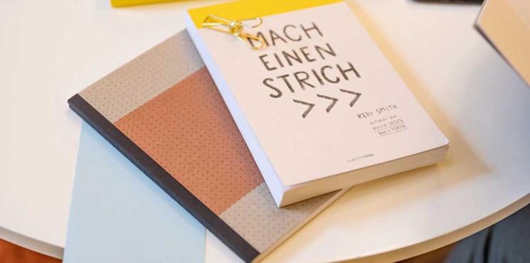 SIGEL Notizbuch in der Farbe Koralle liegt neben einem Block mit der Aufschrift "Mach einen Strich".