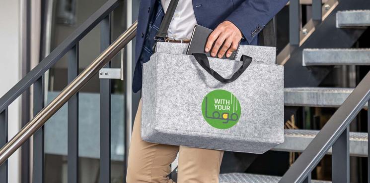 Person trägt eine graue Filztasche mit grünem Logo beim Hinuntergehen einer modernen Treppe