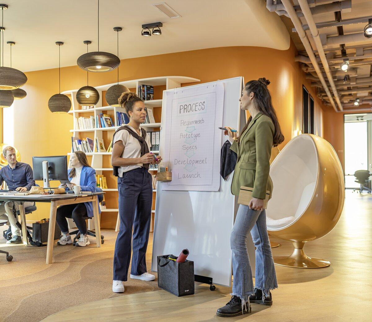 Two people discussing on whiteboard near bookshelf, in office
