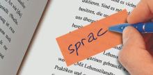 Hand hält blauen Stift, schreibt sprach auf orangenen Zettel über gedrucktem Text