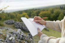 Hände halten Notizbuch und Stift, Hintergrund ist bewaldet