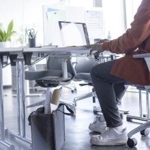 Person am Schreibtisch mit Laptop, Pflanze und Bürobedarf Tasche mit Plänen darunter