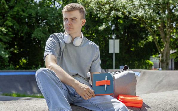 Junger Mann mit Kopfhörern sitzt lässig im Freien und hält Linescape Neo Notizbuch in blau