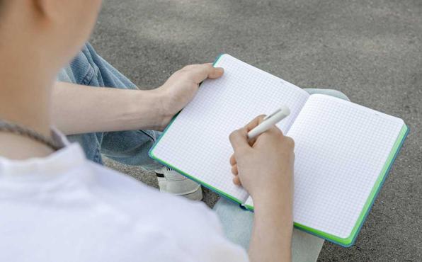 Junger Mann sitzt im Freien, schreibt Notizen in kariertes Linescape Neo Notizbuch