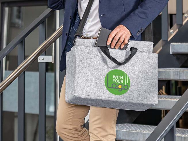 Person trägt eine graue Filztasche mit grünem Logo beim Hinuntergehen einer modernen Treppe