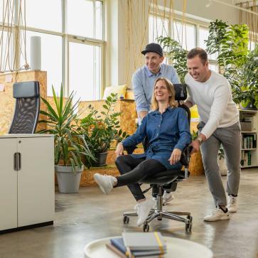 Zwei Männer schubsen Frau auf Schreibtischstuhl an